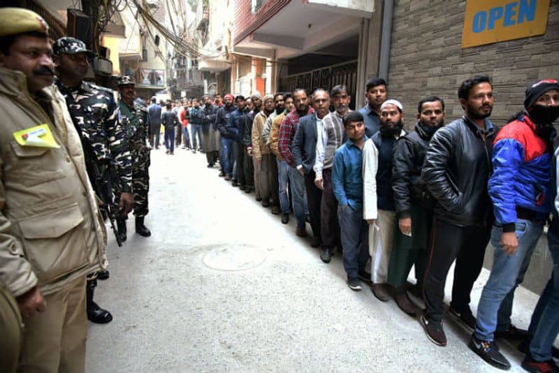 Voters stand in a queue to cast their votes as security personnel stand guard at a polling booth during the Delhi Legislative Assembly elections, at Shaheen Bagh, in New Delhi on Saturday.